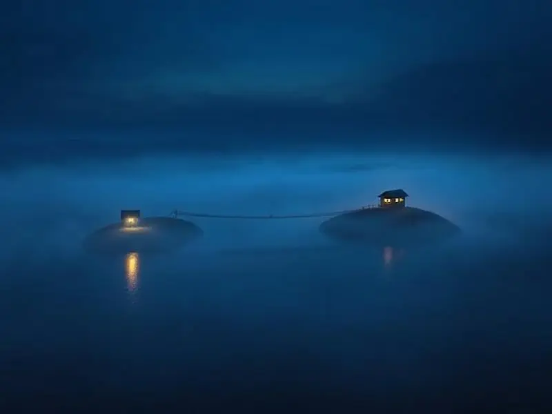 A cinematic photorealistic shot of two separate islands in a calm, misty lake during the blue hour, connected by a delicate, barely visible bridge that seems to float just above the water surface. Each island has a single, warm light glowing from within a small structure, representing two individuals in their separate healing spaces. The scene is captured with a wide-angle lens from an elevated perspective, creating a sense of peaceful separation and respectful distance. The lighting is soft and ethereal, with the blue hour providing natural backlighting while gentle mist diffuses the scene. The bridge appears translucent and magical, suggesting connection that respects boundaries. The color palette features deep blues and purples of twilight, contrasted with warm golden lights from the island dwellings, and silvery highlights on the misty water. Masterpiece, award-winning landscape photography, trending on ArtStation, hyper-detailed mist effects, intricate water reflections, sharp focus, 8K, UHD.
